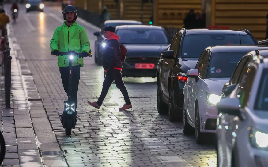 une route avec des voitures, un piétons et un conducteur de trottinette